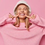 Girl in pink sweater and white hat pointing to her smile with two fingers